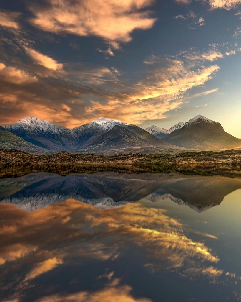 Scenic Scottish Highlands landscape near Dingwall and Grantown‑on‑Spey, with snow‑capped mountains reflected in a tranquil loch at sunrise, under vibrant orange and pink clouds.